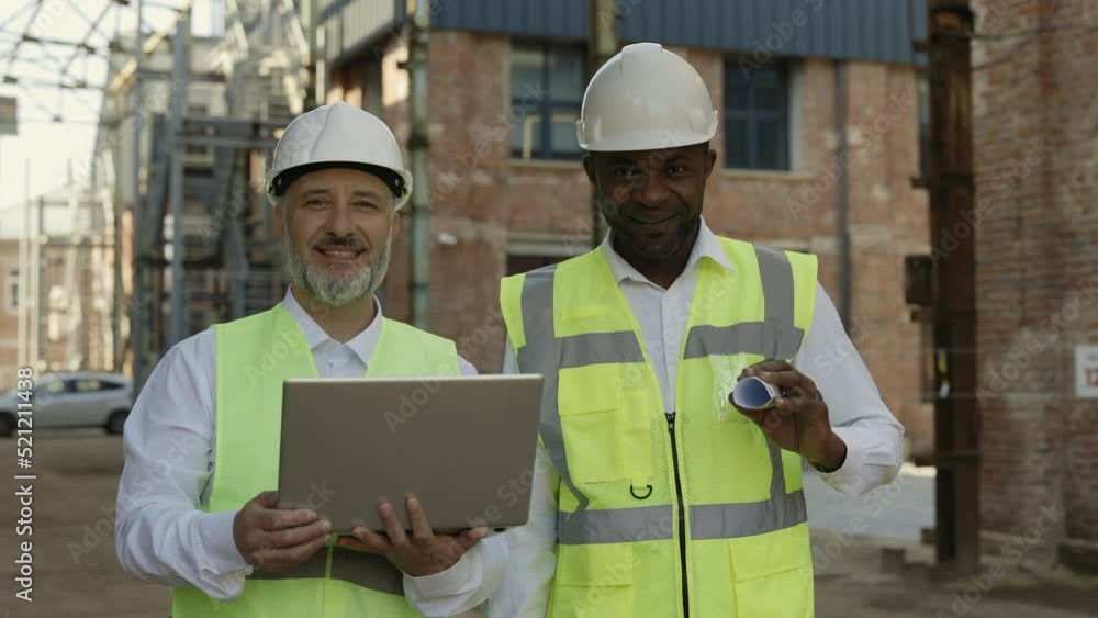 Portrait of smiling construction engineers in safety uniform posing with laptop and blueprints at building site. Multicultural team working together for improving construction process. Portrait of smiling construction engineers in safety uniform posing with laptop and blueprints at building site. Multicultural team working together for improving construction process.