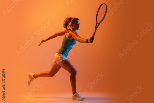 Portrait of sportive woman, professional tennis player training isolated over orange studio background in neon light