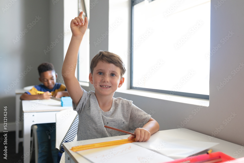 Smiling caucasian elementary schoolboy with hand raised sitting at desk in classroom Smiling caucasian elementary schoolboy with hand raised sitting at desk in classroom