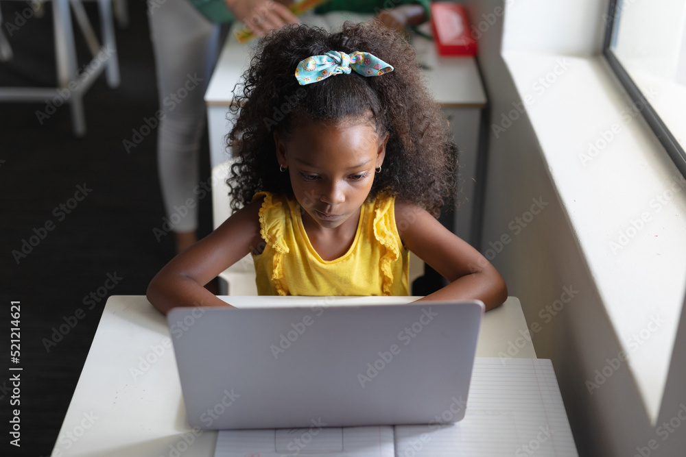 Fototapeta premium High angle view of african american elementary schoolgirl using laptop at desk in classroom