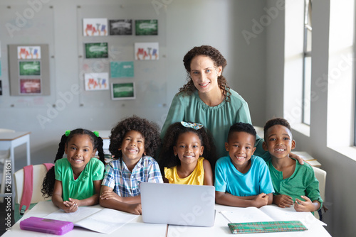 Portrait of smiling caucasian young female teacher with african american elementary students at desk