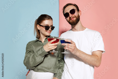 Stylish couple, handsome man and charming girl tasting cocktail isolated over blue-pink background. Fashion, emotions, youth, love, travel, vacation concept