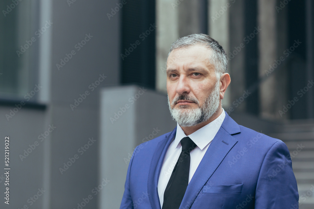 Close-up photo portrait of gray-haired successful businessman boss, man concentrating and thinking looking at camera, businessman owner in business suit outside office Close-up photo portrait of gray-haired successful businessman boss, man concentrating and thinking looking at camera, businessman owner in business suit outside office
