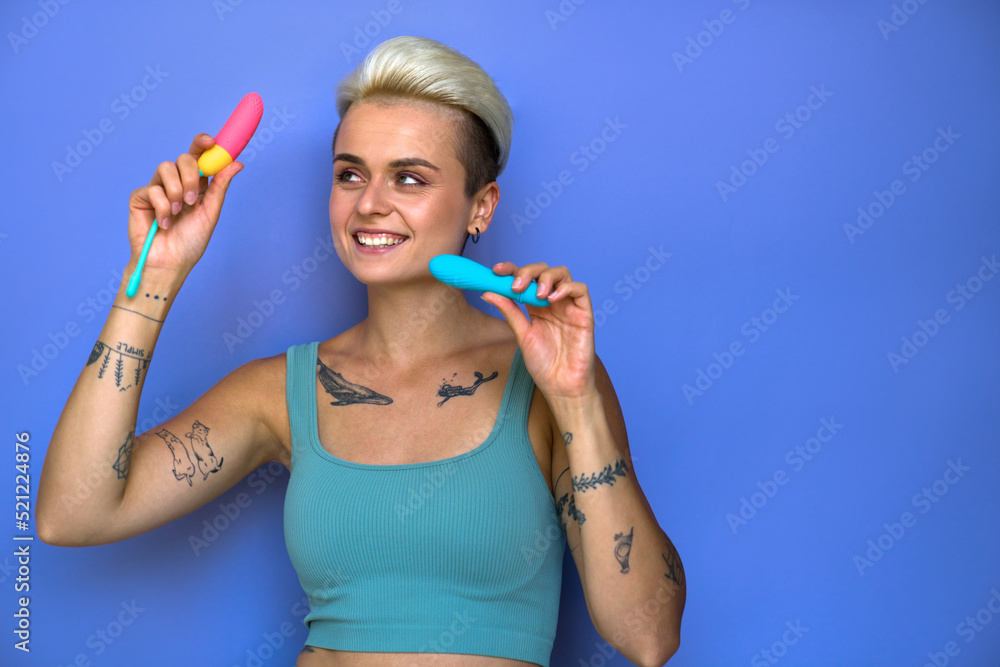 Happy girl in domestic clothes holding two vaginal stimulators over studio background Happy girl in domestic clothes holding two vaginal stimulators over studio background