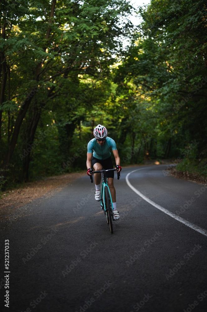 Fototapeta premium Professional female cyclist trains in the woods. Woman riding race bicycle on a forest asphalt road, uphill, front view