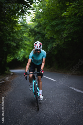 Fototapeta Naklejka Na Ścianę i Meble -  Professional female cyclist trains in the woods. Woman riding race bicycle on a forest asphalt road, uphill, front view. Cycling standing up.