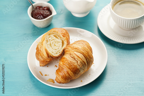 Two french soft croissants on a white plate on a blue wooden table, a cup of black coffee in white sophisticated cup, milk, sugar, jam