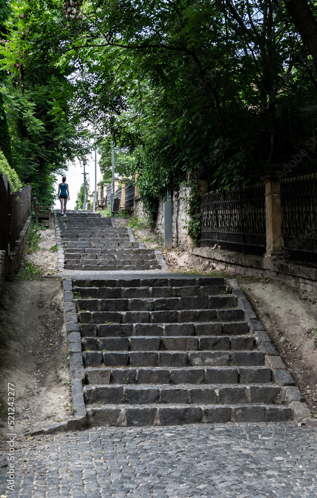 Young sporty woman in sportswear running up steep cobblestone stairs in green alley