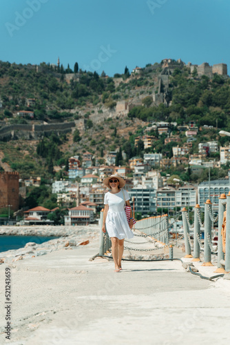 Tourist woman walking on a concrete road along the seashore in a sun hat, white dress and beach bag, resting while traveling. Girl walking on the background of the old city in Alanya. © cinematri