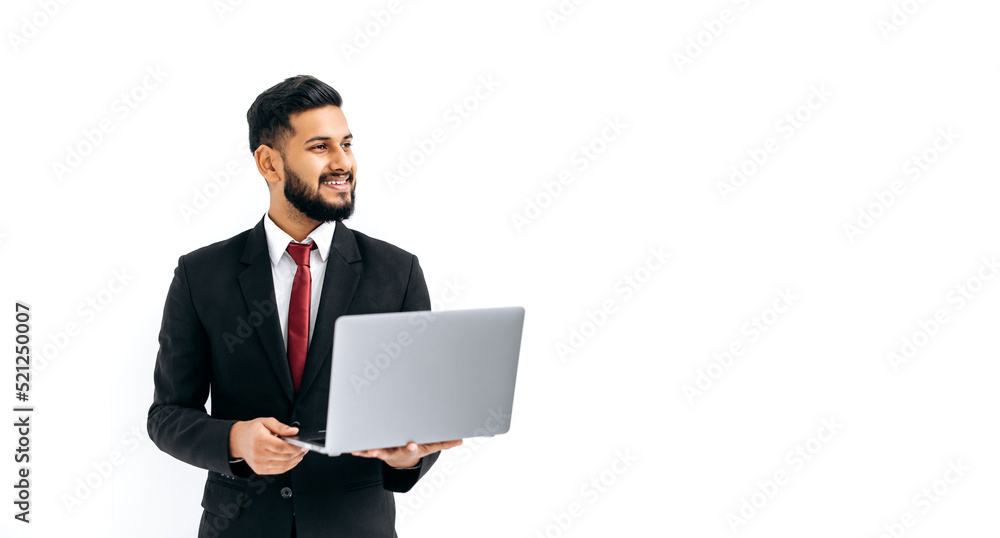 Elegant successful indian or arabian young man in business suit, male executive, holding open laptop in hands, looking aside, stands over isolated white background, smile friendly. Copy-space concept Elegant successful indian or arabian young man in business suit, male executive, holding open laptop in hands, looking aside, stands over isolated white background, smile friendly. Copy-space concept