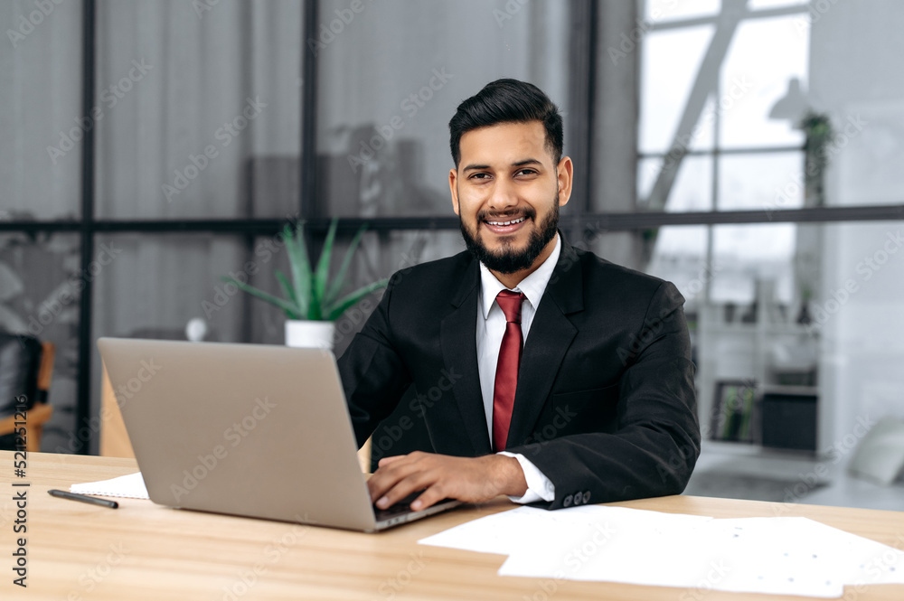 Portrait of handsome confident arabian or indian successful businessman, entrepreneur, lawyer, in a formal suit, sit at work desk with laptop in modern creative office, looks at camera, smile friendly Portrait of handsome confident arabian or indian successful businessman, entrepreneur, lawyer, in a formal suit, sit at work desk with laptop in modern creative office, looks at camera, smile friendly