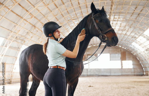 Using brush to comb the hair. A young woman in jockey clothes is preparing for a ride with a horse on a stable photo