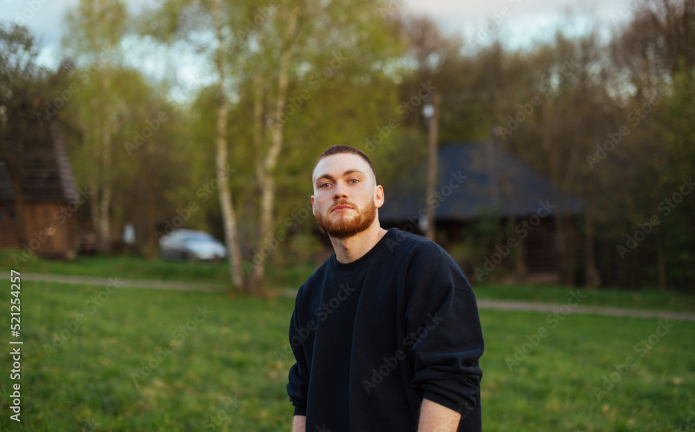 A handsome young man with a beard in casual clothes stands in the park on the background of nature with a serious face A handsome young man with a beard in casual clothes stands in the park on the background of nature with a serious face