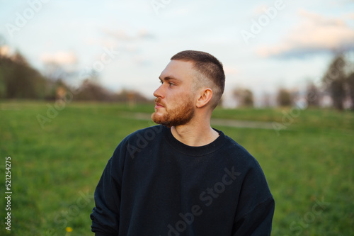 Portrait of a handsome young man with a beard in casual clothes standing in a field in nature and looking away with a serious face.