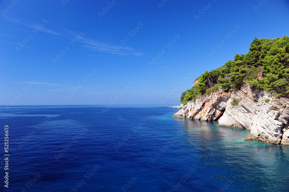 Crystal clear ocean and blue sky in Montenegro, Europe.  White cliffs and green pine trees to the right.  Horizontal photo with large copy space area to the left.