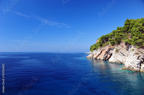 Crystal clear ocean and blue sky in Montenegro, Europe. White cliffs and green pine trees to the right. Horizontal photo with large copy space area to the left.