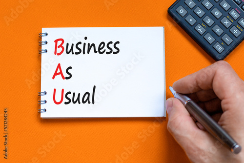 BAU business as usual symbol. Concept words BAU business as usual on white note on a beautiful orange background. Businessman hand. Business and BAU business as usual concept. Copy space.