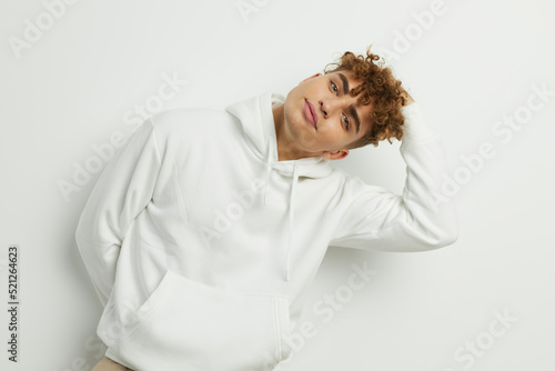 a cute man stands on a white background in a white hoodie with a hood on his head and looks pleasantly into the camera relaxed leaning on the wall