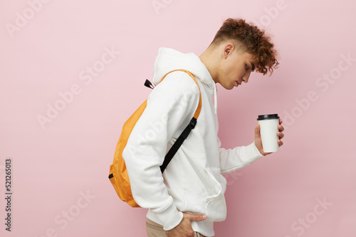 a nice man stands on a pink background in a white hoodie with a backpack on his back, holding a glass with a drink, sideways to the camera looking into the glass