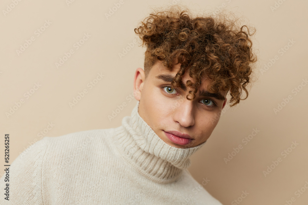 a handsome, attractive man with curly hair stands on a beige background in a light turtleneck pulled over his face a handsome, attractive man with curly hair stands on a beige background in a light turtleneck pulled over his face