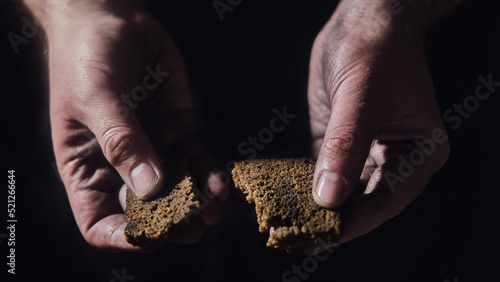 Hungry man holding bread on a black background, hands with food close-up. Dirty hands of a starving poor man on a dark background