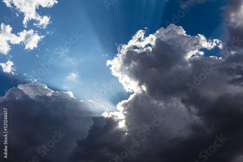 Closeup of cumulus clouds in blue sky. Rays of light emanating from sun behind clouds, creating bright and shadowed beams of light. 