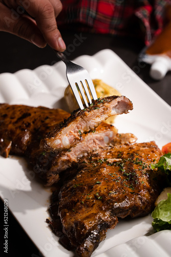 Close up cut in half grilled bbq ribs serve in white square plate and fork pick up a piece of rib on black table