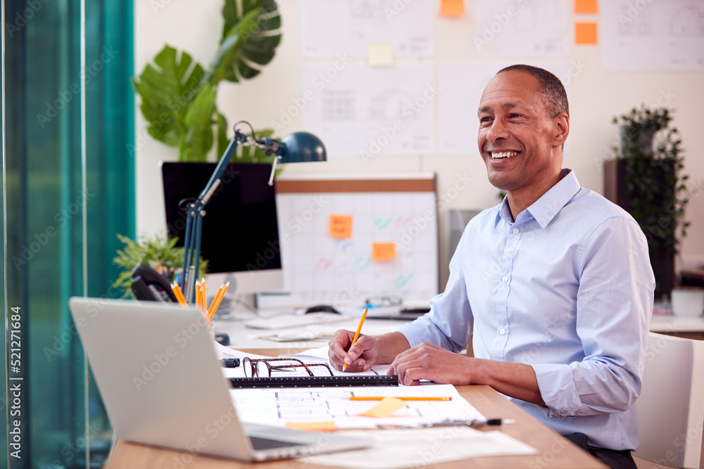 Mature Male Architect Working In Office At Desk Studying Plans For New Building