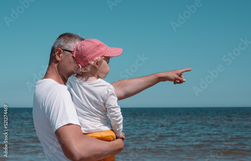 The father holds his daughter in his arms and points to the horizon with his hand.