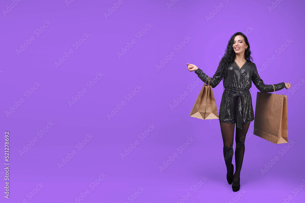 A girl on a shopping trip. A young woman in a black dress with paper bags on a lavender background. A happy smiling girl stands with shopping in her hands. Brown paper bags in hand.