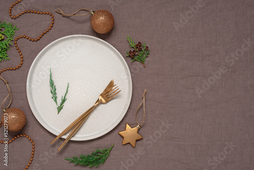 Beige plate with golden cutlery, brown textile background. Christmas minimal table setting with xmas decorations. Top view, flat lay.