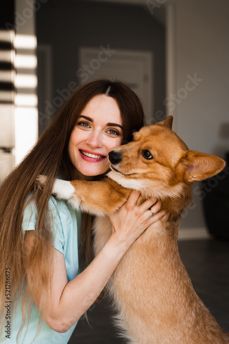 Welsh Corgi Pembroke dog kiss his girl owner at home. Lifestyle with domestic playful pet. Young woman hug lovely Corgi dog and smile.
