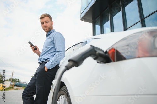 Handsome man in business suit surfing internet on modern smartphone while waiting electric car to charge. © Serhii