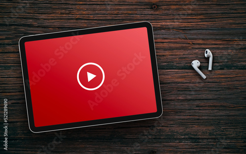 Play button on red screen of tablet and wireless earphones on dark wooden surface