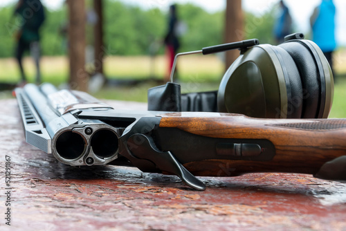 A sports double-barreled shotgun and headphones for shooting lie on a wooden table against the background of the shooting range. photo