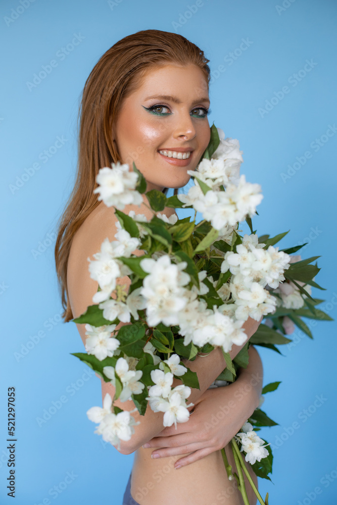 Photo closeup of happy smiling topless woman with bright evening makeup covering with bouquet of fresh white flowers on blue background. Advertising shooting, professional makeup and beauty products