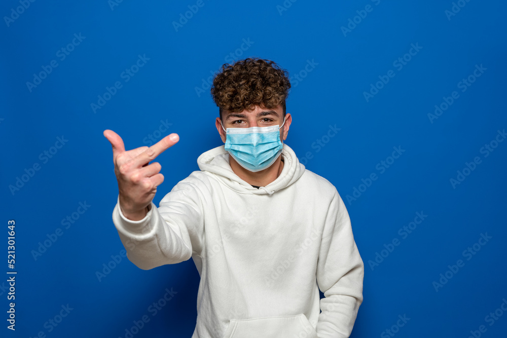 Portrait of angry young man in surgical medical mask and white sweatshirt standing with thumb up and looking at camera with serious or poker face. Indoor studio shot on blue background
