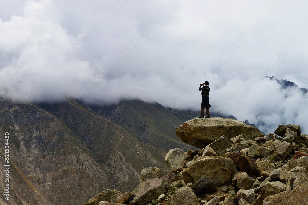 A man taking a photo of the valley engulfed in clouds, while standing on rocks in Hispar valley of Gilgit Baltistan region of Pakistan. 