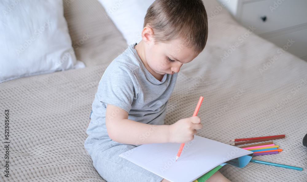 A small caucasian boy in a gray T-shirt and shorts on the bed draws with colored pencils in a sketch A small caucasian boy in a gray T-shirt and shorts on the bed draws with colored pencils in a sketch