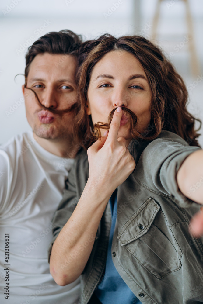 Happy newlyweds play and pose making mustaches with curly brunette hair and kiss smiling broadly for selfies via smartphone closeup Happy newlyweds play and pose making mustaches with curly brunette hair and kiss smiling broadly for selfies via smartphone closeup