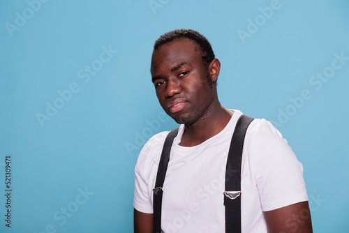 Smiling young adult person wearing white tshirt and suspenders posing alone at camera. Handsome cool looking guy standing on blue background while looking at camera. Studio shot