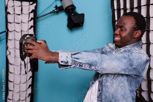 Professional studio producer taking selfie photo with DSLR camera while standing beside modern photo equipment on blue background. Young photographer taking picture of himself while smiling heartily.