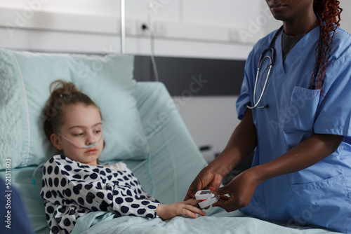Ill kid sitting in hospital pediatric ward while medical staff monitoring health condition using oximeter. Nurse measuring oxygen levels of hospitalized sick little girl resting on patient bed