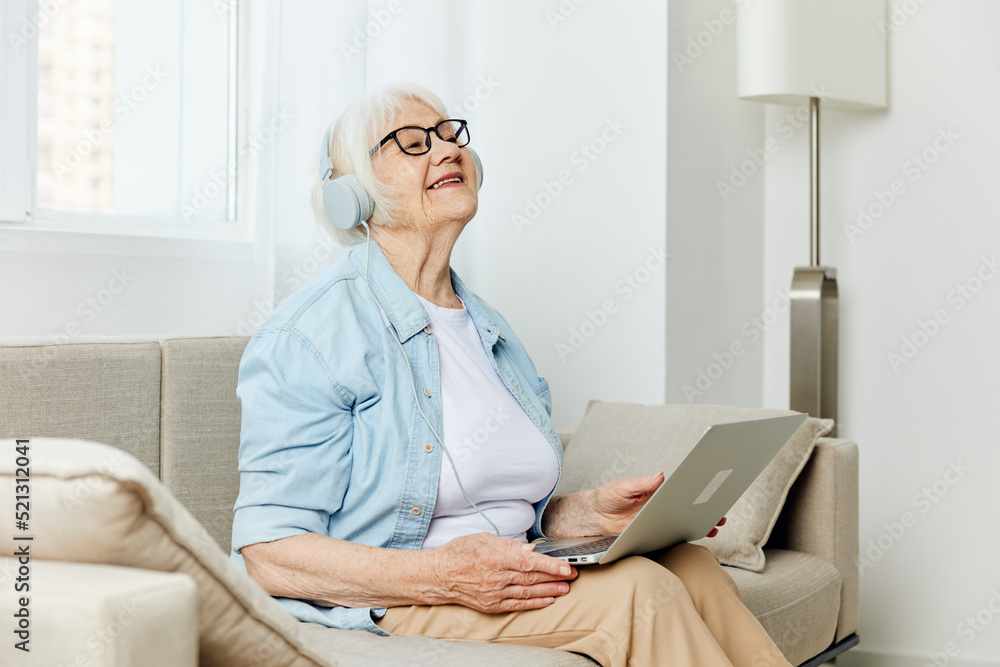a happy, emotional elderly woman with gray hair is sitting at home on a cozy sofa with headphones on her head and happily smiling holding a laptop on her lap a happy, emotional elderly woman with gray hair is sitting at home on a cozy sofa with headphones on her head and happily smiling holding a laptop on her lap