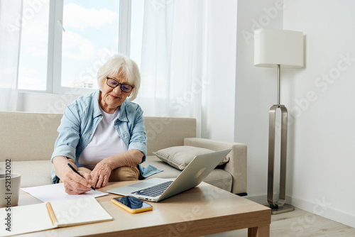 an elderly woman in stylish clothes is sitting on a sofa in a comfortable bright apartment and working on a laptop makes notes in a notebook. The concept of working from home