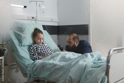 Ill little girl sleeping on patient bed inside hospital pediatrics ward while father sitting beside her. Sick kid under treatment resting inside healthcare clinic while parent waiting her to wake up.