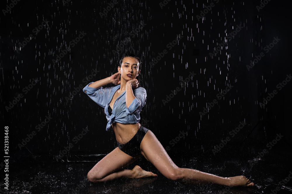 Beautiful girl in a black swimsuit and shirt sits in the rain, enjoys the drops of water. Looks into the camera. Water splash on black background. Aqua studio