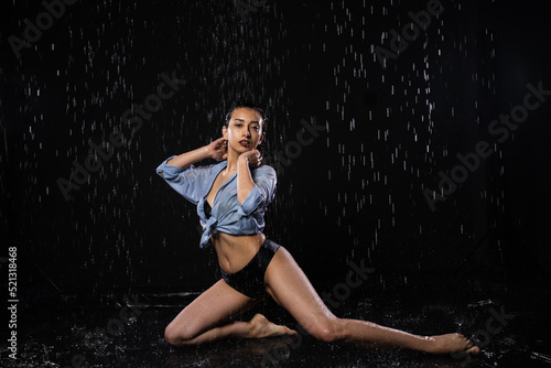 Beautiful girl in a black swimsuit and shirt sits in the rain  enjoys the drops of water. Looks into the camera. Water splash on black background. Aqua studio