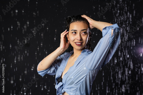 Portrait of a beautiful girl in a black swimsuit and shirt. Standing in the rain Smiling and holding her hair with her hands. Water splash on black background. Aqua studio