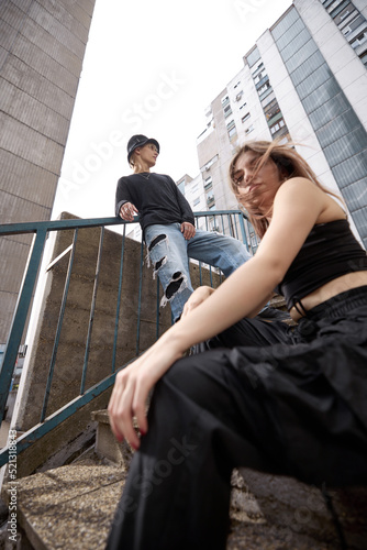 Low angle view of a teenage couple poses in the urban exterior on the stairs surrounded by skyscrapers and buildings.